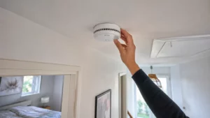 Alarme incendie maison : ce que dit vraiment la loi Installation d’un détecteur de fumée DAAF au plafond d’un couloir dans un logement résidentiel français, avec une main en train de fixer l’appareil conformément aux obligations de sécurité incendie.