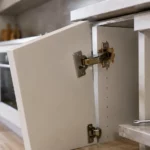 Charnière invisible de porte de placard de cuisine partiellement arrachée dans un panneau aggloméré mélaminé, avec vis sortie du support et porte affaissée avant réparation.