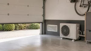 clim réversible dans un garage : ce que dit la physique Unité extérieure de climatisation réversible installée dans un garage bien ventilé avec une porte sectionnelle entrouverte et une grille d'aération murale favorisant le renouvellement de l'air.